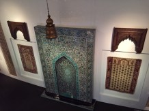 Items for mosques from around the world.  In the center is a tiled Mirhab, prayer rugs on either side, and a lamp hanging from above.