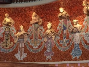 On both sides of the stage are figures with protruding heads and instruments in relief and mosaic bodies. There are nine female figures on each side.